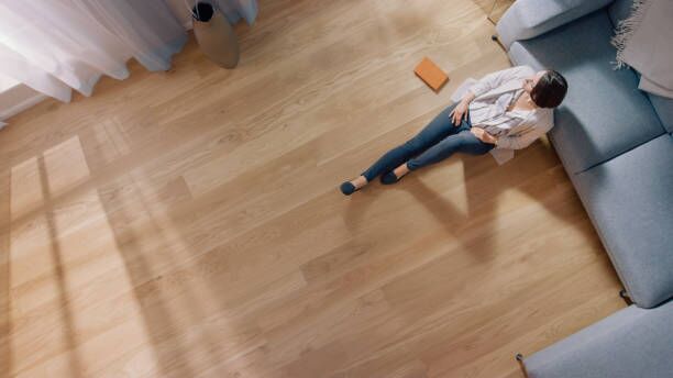 Young Woman is Sitting on a Floor and Looking out of the Window after Reading a Book. Cozy Living Room with Modern Interior, Grey Sofa and Wooden Flooring. Top View Camera Shot. Young Woman is Sitting on a Floor and Looking out of the Window after Reading a Book. Cozy Living Room with Modern Interior, Grey Sofa and Wooden Flooring. Top View Camera Shot. Beautiful home stock pictures, royalty-free photos & images