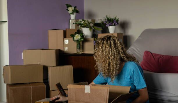 Beautiful young woman moving to new apartment, sitting down while opening the boxes, arranging her stuff. Stock photography of a beautiful young woman with curly hair moving in to her new appartment, opening boxes, arranging her stuff and flowers. long distance movers stock pictures, royalty-free photos & images