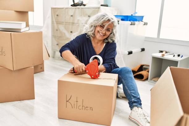 Middle age grey-haired woman smiling happy packing kitchen cardboard box at new home. Middle age grey-haired woman smiling happy packing kitchen cardboard box at new home. Apartment packing stock pictures, royalty-free photos & images