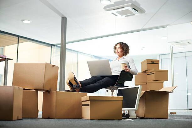 new business start up dreamer A business owner sits amongst her moving in boxes , waiting for the removal team to bring the desks . She is on his laptop keeping business going during the transition . She looks off camera and seems confident of the new office space. moving company stock pictures, royalty-free photos & images
