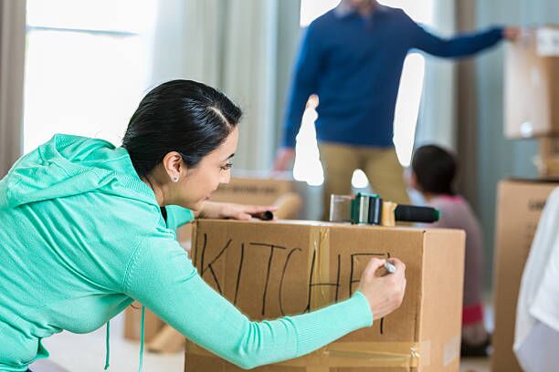 Woman labels box with kitchen supplies Mid adult Hispanic woman writes ‘kitchen’ with a marker. She has just packed up some of the kitchen supplies as she and her family prepare to move. Her husband and daughter are working in the background. labeling boxes stock pictures, royalty-free photos & images